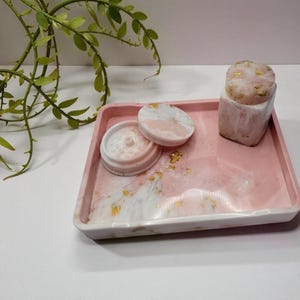 May include: A pink resin tray with a white and pink marbled design, holding a grinder, a lid, and a small container. Gold flakes are scattered throughout. A green plant is in the background.