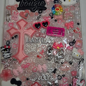 May include: A clear tablet case decorated with various pink and white charms, including Hello Kitty figures, a large pink cross, a glittery pink guitar, bows, hearts, and text reading a lil bougie, LAY LAY, and Queen, along with a pink Lonely Hearts Club ticket.
