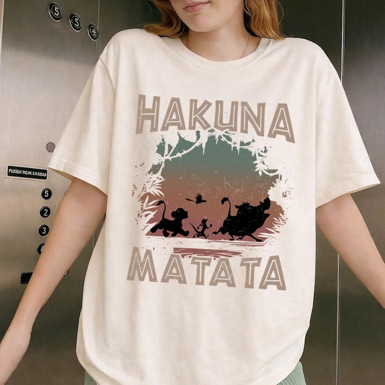 Puede incluir: Camiseta color crema con la frase "HAKUNA MATATA" en letras marrones. El dise&ntilde;o incluye siluetas de Simba, Tim&oacute;n, Pumba y un p&aacute;jaro dentro de un marco con tem&aacute;tica de jungla. Es una prenda informal.