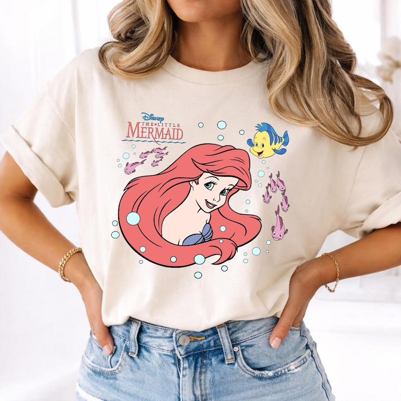 May include: Cream-colored t-shirt featuring a graphic of Ariel from Disney's The Little Mermaid. Ariel has long red hair and is surrounded by bubbles and fish. The shirt also displays the movie's title in a stylized font.