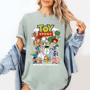 Op de afbeelding: Lichtgroen T-shirt met een kleurrijke afbeelding van personages uit de animatiefilm "Toy Story". Het ontwerp bevat de woorden "TOY STORY" en de zin "YOU'VE GOT A FRIEND IN ME" onder de personages.