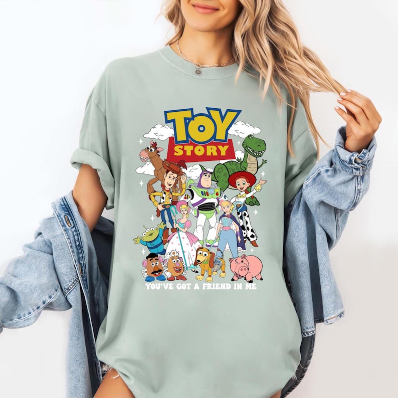 Op de afbeelding: Lichtgroen T-shirt met een kleurrijke afbeelding van personages uit de animatiefilm "Toy Story". Het ontwerp bevat de woorden "TOY STORY" en de zin "YOU'VE GOT A FRIEND IN ME" onder de personages.