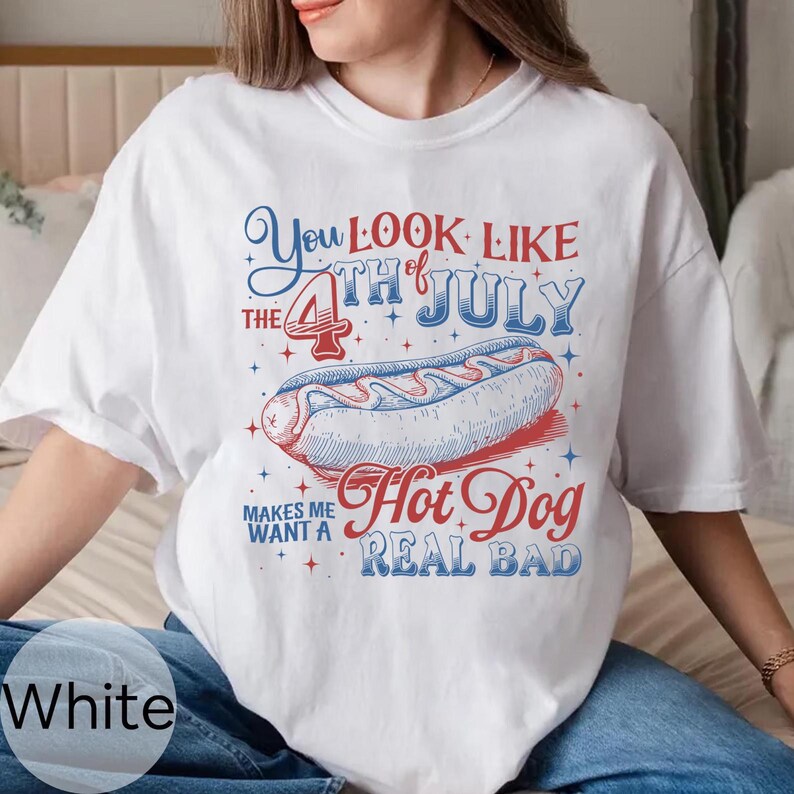 Op de afbeelding: Wit T-shirt met een afbeelding van een hotdog en de tekst "You Look Like The 4th of July Hot Dog Makes Me Want A Real Bad" in rood en blauw. Het ontwerp is gecentreerd op de voorkant.