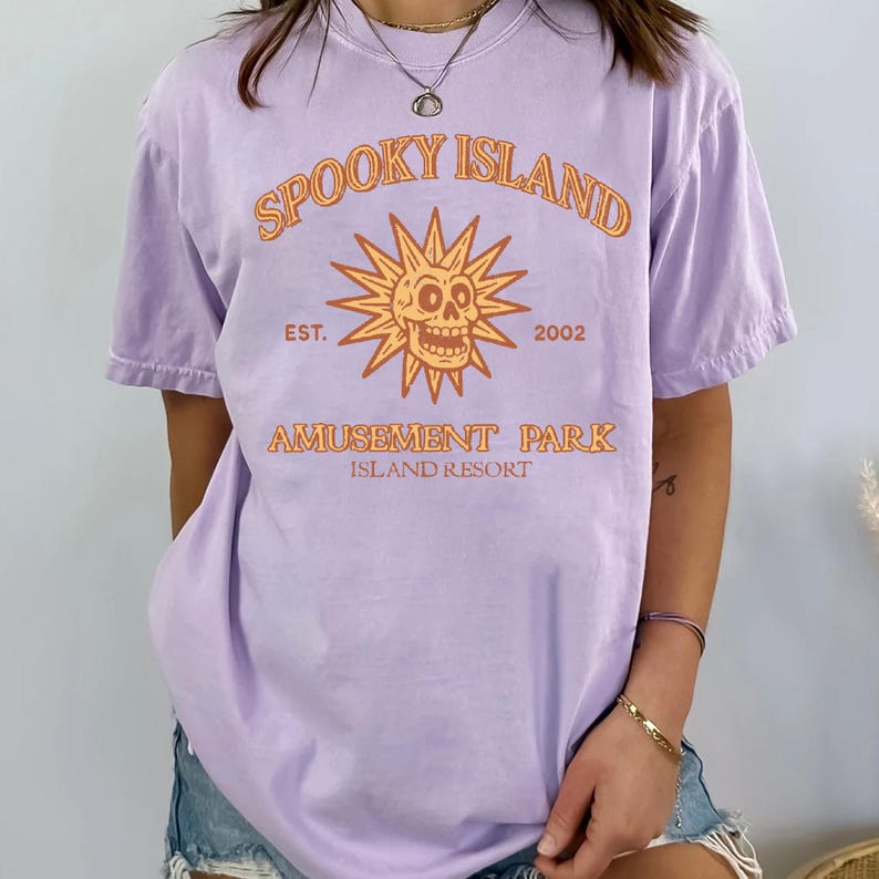 May include: A lilac-colored t-shirt with the text "Spooky Island Amusement Park Island Resort" in orange. The design includes a sun with a skull face and the year "2002". The shirt has short sleeves and a relaxed fit.