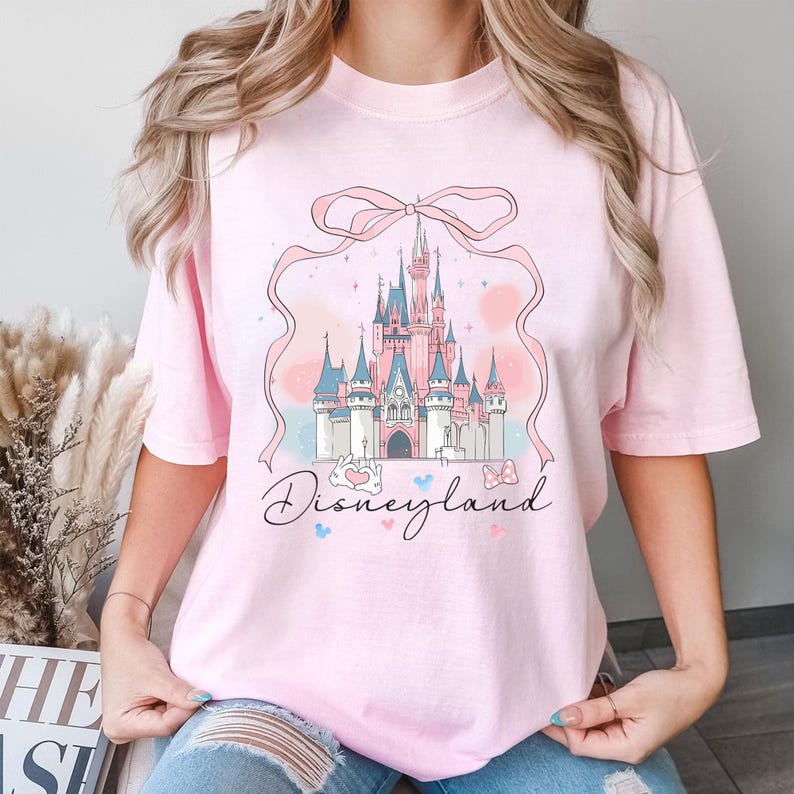 May include: A light pink t-shirt featuring a whimsical illustration of a fairytale castle with pink and blue accents, a pink ribbon, and the word "Disneyland" in script. The design includes hearts and bows.