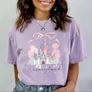 May include: A lilac-colored t-shirt featuring a pink castle graphic with a bow and the word "Disneyland" in script. The castle has pink and blue turrets and is set against a pastel background. Butterflies and hearts accent the design.