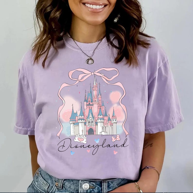 May include: A lilac-colored t-shirt featuring a pink castle graphic with a bow and the word "Disneyland" in script. The castle has pink and blue turrets and is set against a pastel background. Butterflies and hearts accent the design.