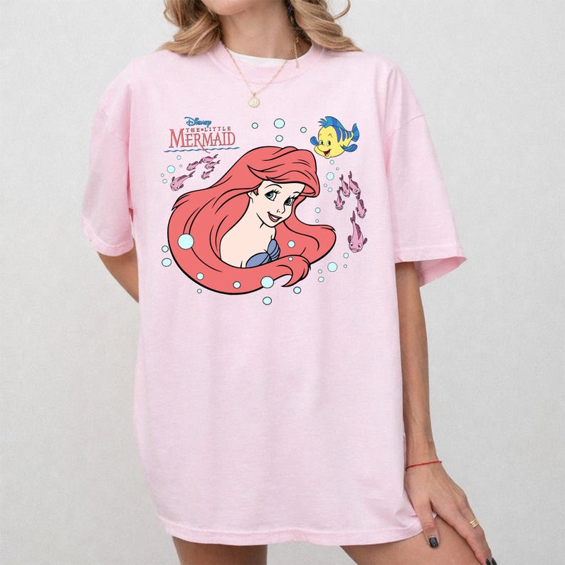 May include: A light pink t-shirt featuring a graphic of Ariel from Disney's The Little Mermaid. The design includes Ariel with red hair, a blue seashell top, and Flounder. The shirt has the words "Disney The Little Mermaid" printed above the graphic.