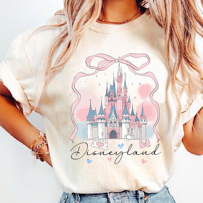 May include: Cream-colored t-shirt featuring a pink and blue illustration of a castle with a pink ribbon and the word "Disneyland" in script. The castle has blue and pink turrets and is set against a pastel background.
