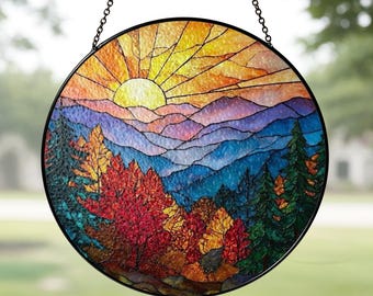 May include: Round stained glass window art depicting a mountain landscape with a setting sun. The artwork features vibrant colors, including orange, yellow, red, blue, and green. The piece is framed in black and hangs from a chain.
