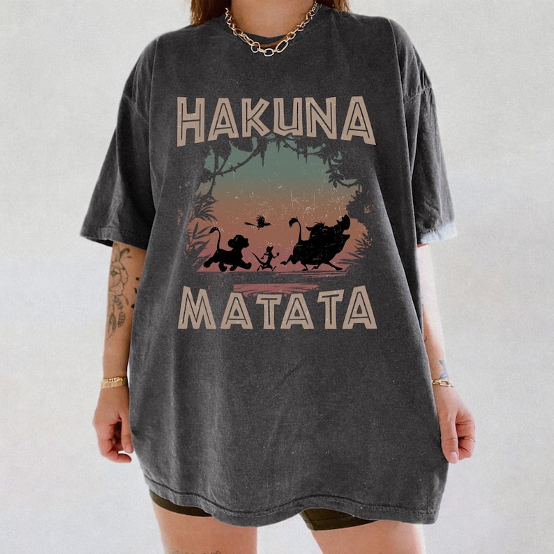 Puede incluir: Camiseta gris oscuro con las palabras "HAKUNA MATATA" en fuente beige. La camiseta presenta un gr&aacute;fico colorido de siluetas de personajes de El Rey Le&oacute;n. La camiseta es de corte holgado.