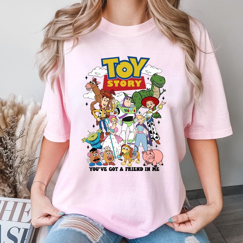 Op de afbeelding: Lichtroze t-shirt met een kleurrijke afbeelding van personages uit de film "Toy Story". Het ontwerp bevat het logo van de film en de zin "You've Got a Friend in Me."