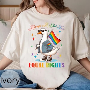 May include: Ivory-colored t-shirt featuring a cartoon goose wearing sunglasses, a denim jacket, and sneakers, holding a rainbow flag. The text "Always A Slut For" and "EQUAL RIGHTS" are printed in colorful fonts. The design includes star accents.