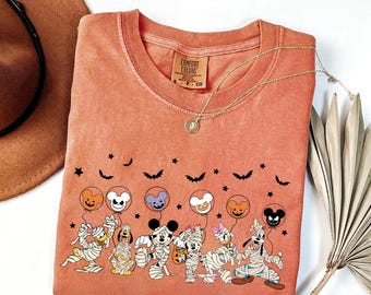 May include: Orange Comfort Colors t-shirt featuring a Halloween-themed graphic. The graphic includes cartoon characters dressed as mummies, holding balloons with character faces. Bats and stars are also included in the design. A gold necklace with a pendant is draped over the shirt.
