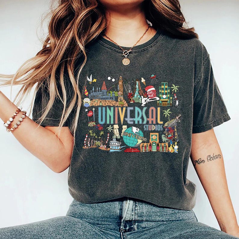 May include: Dark gray t-shirt featuring a colorful graphic of Universal Studios attractions. The design includes the text "UNIVERSAL STUDIOS" in rainbow colors, with a globe and various theme park icons.