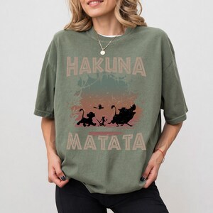 Puede incluir: Camiseta verde oliva con las palabras "HAKUNA MATATA" en una fuente marr&oacute;n claro. La camiseta tiene un gr&aacute;fico de siluetas de Simba, Tim&oacute;n, Pumba y p&aacute;jaros. La camiseta es de corte holgado.