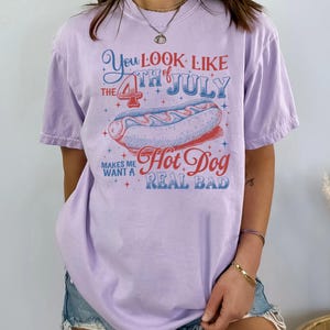 Op de afbeelding: Lichtpaarse t-shirt met een afbeelding van een hotdog en de tekst "You Look Like The 4th of July Makes Me Want A Hot Dog Real Bad" in rood en blauw. Ideaal voor de viering van 4 juli.
