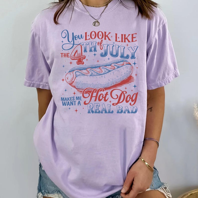 Op de afbeelding: Lichtpaarse t-shirt met een afbeelding van een hotdog en de tekst "You Look Like The 4th of July Makes Me Want A Hot Dog Real Bad" in rood en blauw. Ideaal voor de viering van 4 juli.