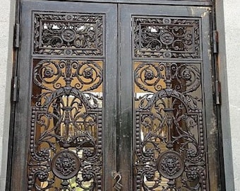 May include: A set of double doors made of black metal featuring intricate scrollwork, floral motifs, and circular medallions with lion head reliefs. The doors include a rectangular transom window above, also decorated with matching metal scrollwork.