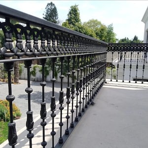 May include: Black wrought iron balcony railing with ornate decorative elements. The railing features vertical balusters and a top rail with a repeating pattern. The background includes greenery and a building.
