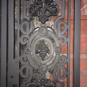 May include: A rectangular black metal door panel featuring an intricate cast iron design. The pattern includes symmetrical scrollwork, floral motifs, and a central oval frame containing a decorative urn element. The frame has a keyhole cutout.