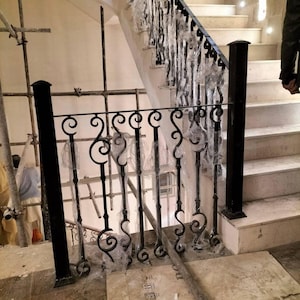 Può includere: Ringhiera per scale in ferro battuto nero con volute decorative e barre verticali. La ringhiera è installata accanto a una scala con gradini bianchi e un corrimano nero. La ringhiera è avvolta in plastica protettiva.