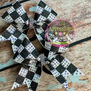May include: Two cheerleading bows with a black and white checkered pattern and sparkling rhinestones. The bows are embellished with a rhinestone center and are set against a wooden background. The bows have the text "Mini Xpressions".