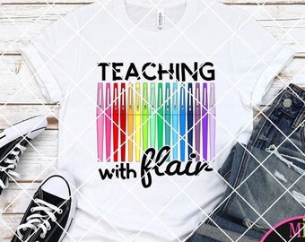 May include: White t-shirt with the text "Teaching with flair" and a rainbow of colorful markers. The shirt is on a wooden background with a pair of black sneakers and blue jeans.