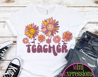 May include: White t-shirt with a pink, orange, and yellow sunflower design and the word "TEACHER" in a retro font.