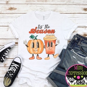 May include: White t-shirt with a graphic of a pumpkin and a coffee cup with the text "Tis' the Season". The shirt is paired with blue jeans and black sneakers.