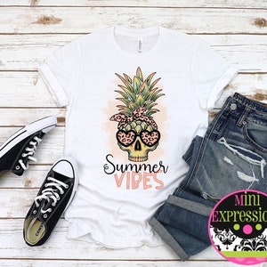 May include: White t-shirt with a pineapple skull graphic and the text "Summer Vibes". The pineapple skull is wearing sunglasses and a bandana. The t-shirt is laying on a wooden surface with a pair of black sneakers and blue jeans.