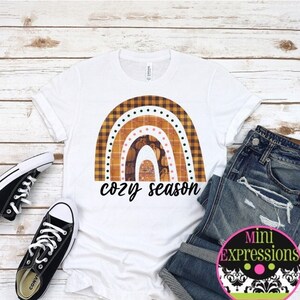May include: White t-shirt with a rainbow design made of plaid fabric and the text "cozy season". The rainbow is in shades of brown, orange, and yellow. The t-shirt is laid out on a wooden surface with a pair of black sneakers and blue jeans.