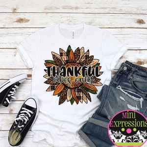 May include: White t-shirt with a sunflower design and the text "THANKFUL vibes only." The sunflower has plaid and fall colors. Black canvas sneakers and blue jeans are also in the image.
