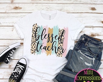 May include: White t-shirt with the words "Blessed Teacher" in a script font, with colorful brushstroke accents. The shirt is paired with blue jeans and black sneakers on a light wood background.
