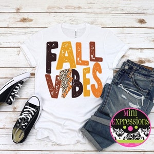 May include: White t-shirt with the text "Fall Vibes" in orange, brown, and yellow letters with a leopard print design. The shirt is on a wooden background with a pair of black sneakers and blue jeans.