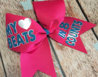 May include: A pink cheer bow with a silver heart and blue glitter text that reads "My Beats In 8 Counts".