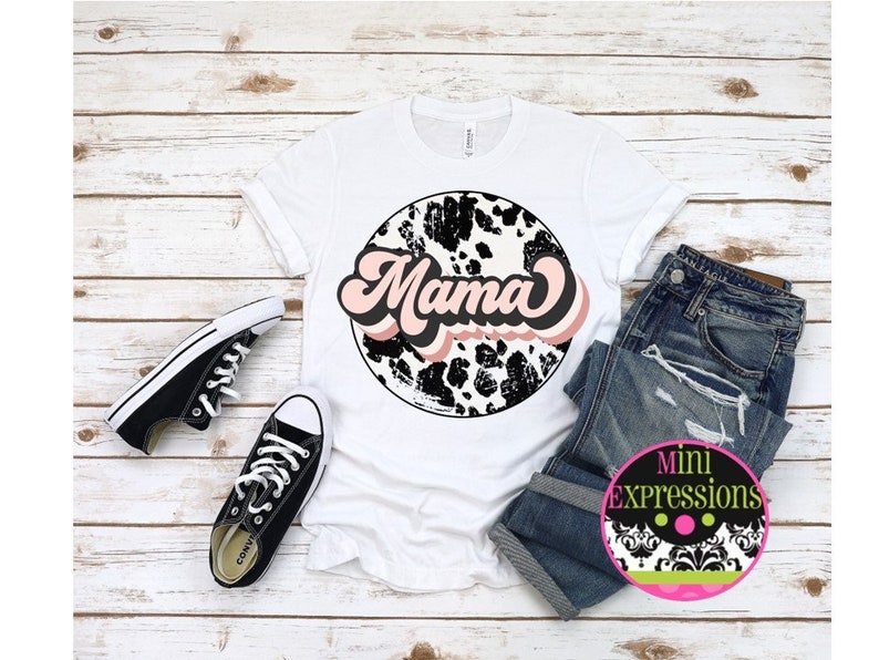 K&ouml;nnte beinhalten: Wei&szlig;es T-Shirt mit einem schwarzen und wei&szlig;en Kuhfleckenkreis mit dem Wort "Mama" in rosa und schwarzen Buchstaben.