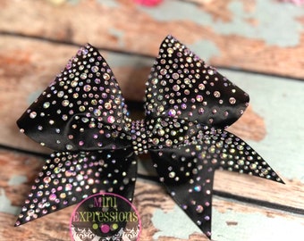 May include: A black cheer bow with iridescent rhinestones. The bow is on a wooden surface. The text "Mini Expressions" is visible on the bow.
