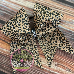 May include: A large gold and black cheetah print cheer bow with rhinestones. The bow is tied with a black elastic band.