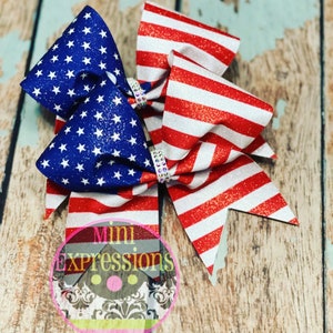 May include: Two red, white, and blue glitter cheer bows with rhinestones. The bows are in the shape of a traditional bow with two loops and a center knot. The bows are on a wooden background.