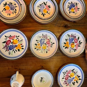 May include: A collection of ceramic dinnerware featuring white plates, bowls, and a creamer with hand-painted fruit and floral motifs in blue, yellow, pink, and green. Each piece is bordered by a dark green rim and a blue cross-hatched pattern.