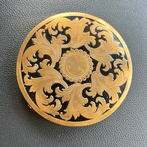 May include: A round, antique compact with a gold-colored metal frame and a black center. The compact features an ornate, symmetrical design of swirling leaves and a central circular detail. The compact is closed.