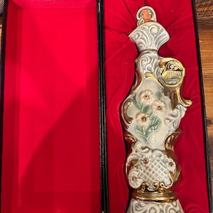 May include: A white porcelain decanter with gold accents and floral designs, in a red velvet lined box. The decanter is shaped like a vase with a wide base and a narrow neck. The box is black with gold trim.