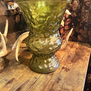 May include: A green glass vase with a textured surface. The vase has a wide top and a bulbous base, sitting on a wooden surface. The vase is empty and appears to be vintage.