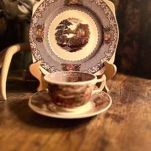 May include: A vintage tea set featuring a square plate and a teacup with a saucer. The set has a brown and white color scheme with a landscape design in the center of the plate and cup. The plate is supported by a wooden stand.