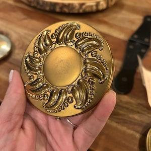 May include: A gold-toned compact mirror with an ornate design featuring a central circle surrounded by a decorative border of swirling leaves and beads.
