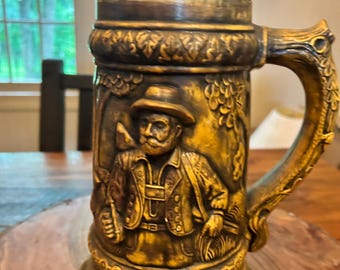May include: A vintage ceramic beer stein, predominantly brown and gold, with a detailed relief of a man in traditional attire. It features a hinged lid and an ornate handle. The design includes a figure on the lid and a detailed scene on the body.