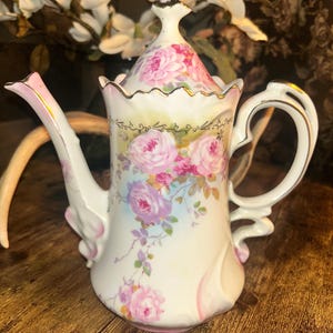 May include: A white porcelain teapot with a lid, adorned with pink roses and gold trim. The teapot features a curved spout and handle, with additional pink accents. The background includes white flowers and a wooden surface.
