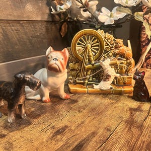 May include: A collection of ceramic figurines, including a terrier, bulldog, cat, and dog. A decorative spinning wheel scene is also present. The figurines are in various colors, including brown, white, and black.