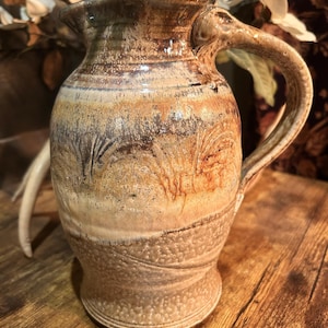 May include: A handcrafted ceramic pitcher with a unique, textured surface. The pitcher features a warm color palette of browns, tans, and creams, with a curved handle and a slightly flared spout. The design includes subtle, organic patterns.